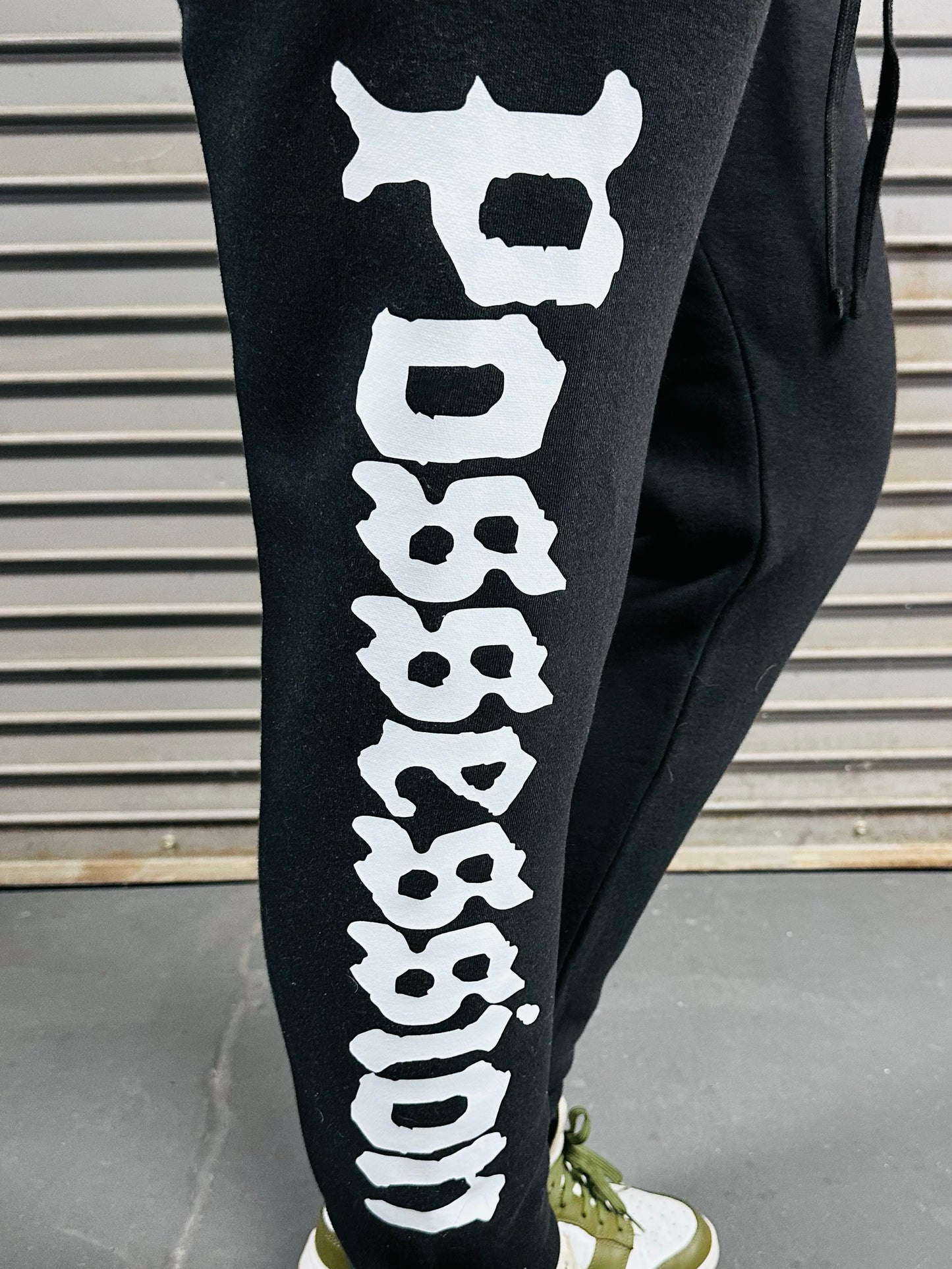 Black pants with white 'Possession' text worn by a person, against a metallic background.