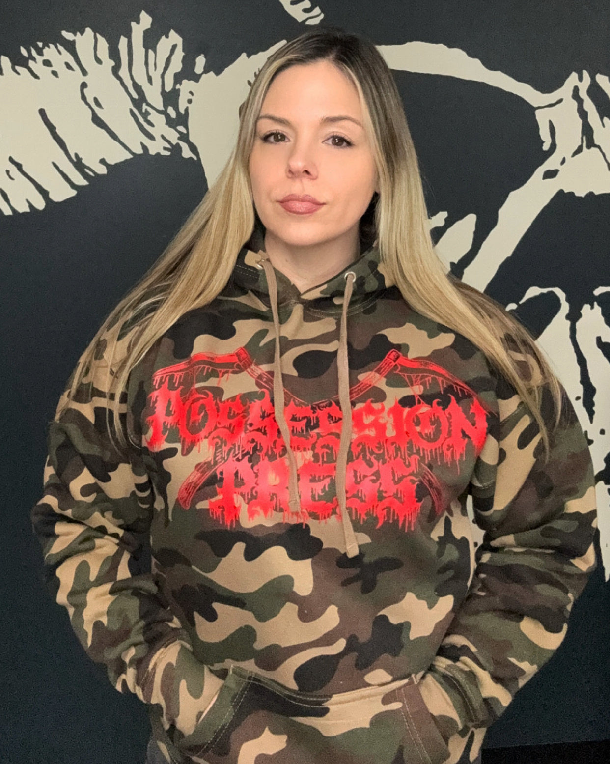 Woman wearing a camouflage hoodie with red "Possession Press" text against a black background with white designs.