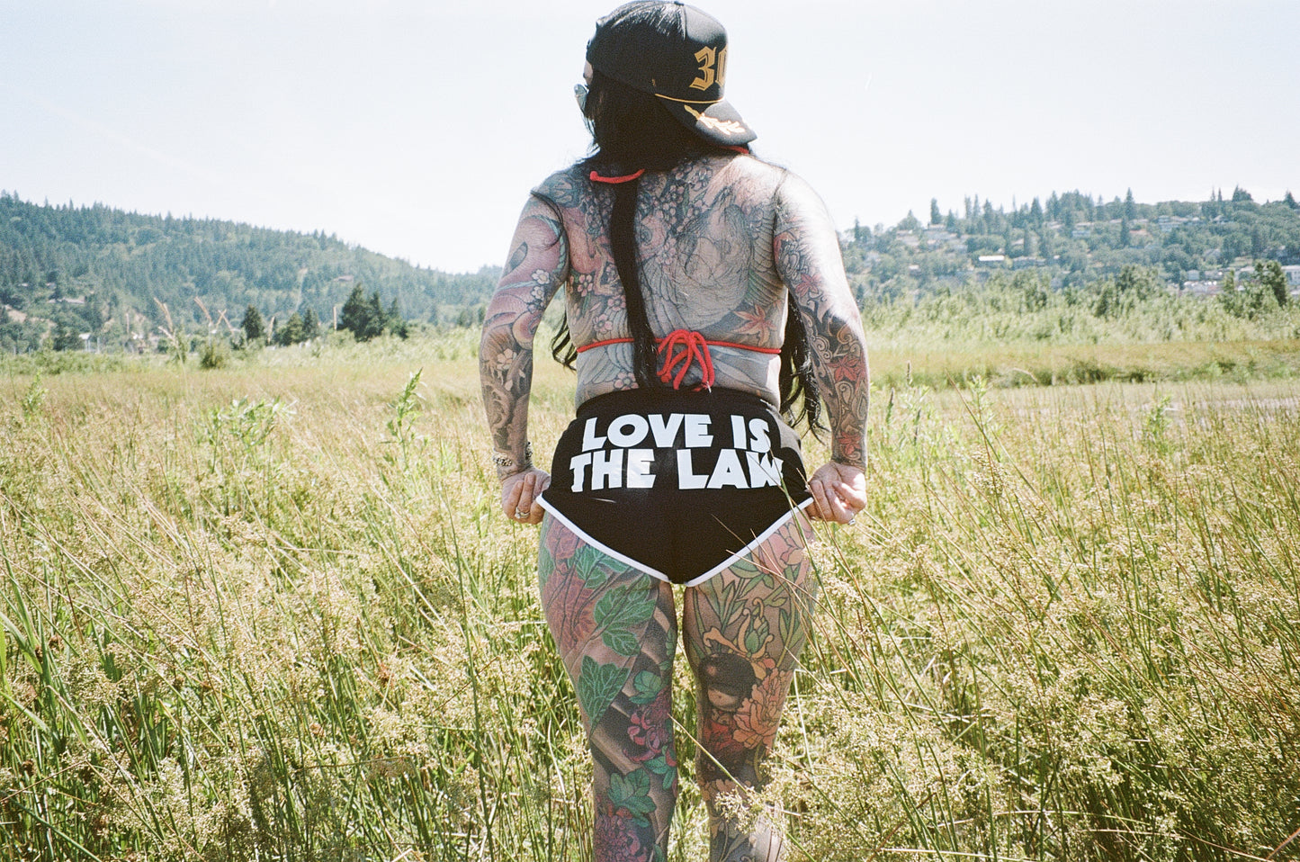 tattooed woman standing in a field with "love is the law" shorts on