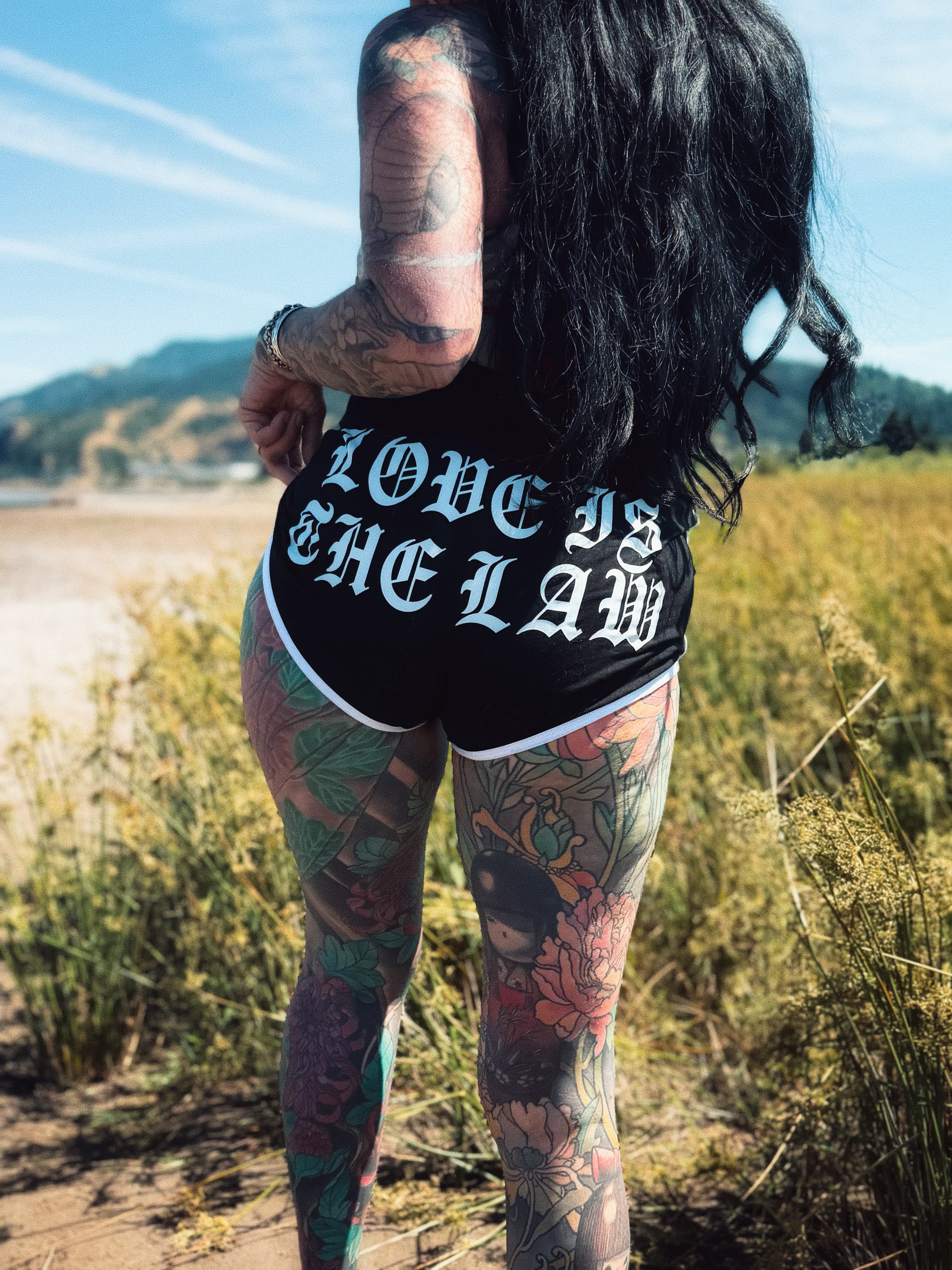 tattooed woman standing in a field with "love is the law" shorts on