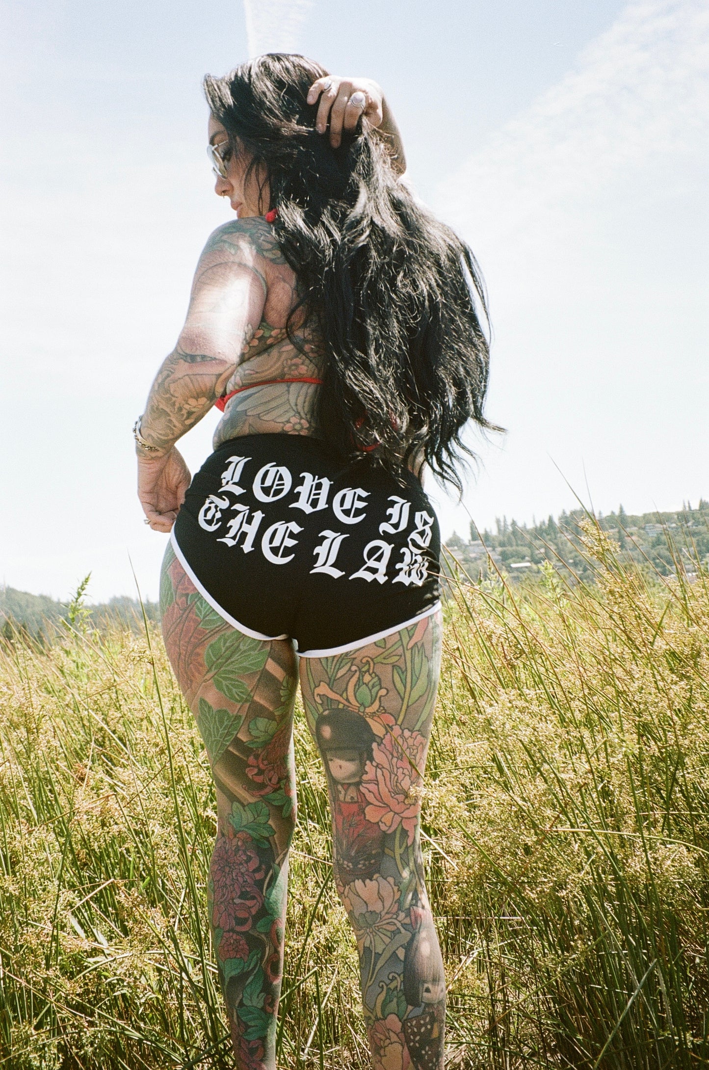 tattooed woman standing in a field with "love is the law" shorts on