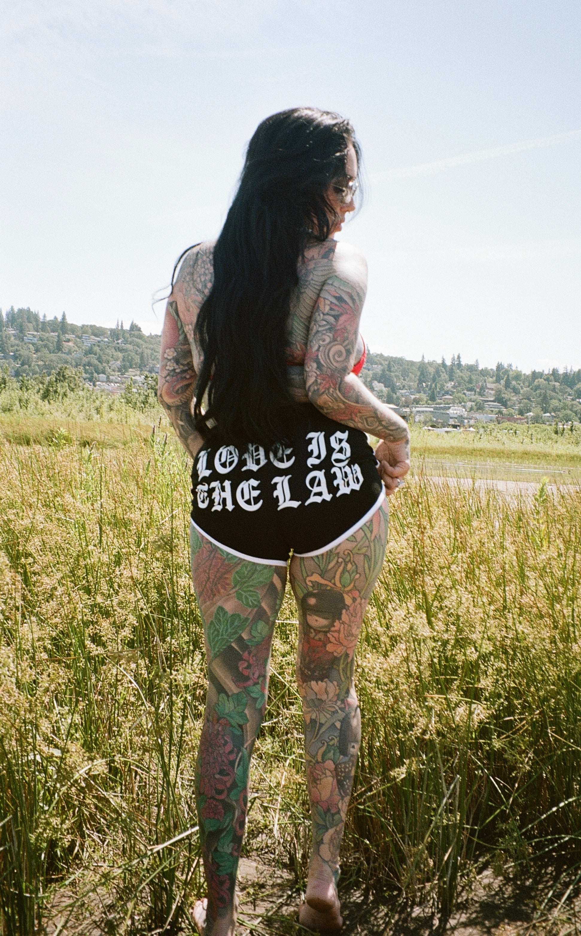 tattooed woman standing in a field with "love is the law" shorts on