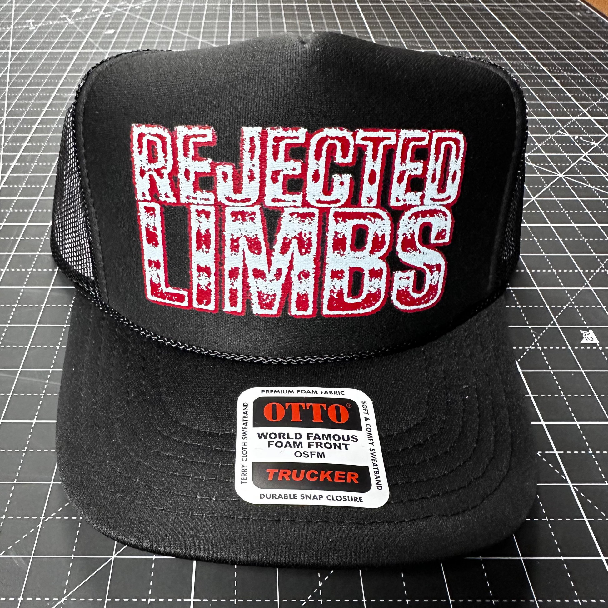 Black trucker hat with 'Rejected Limbs' text on a cutting mat