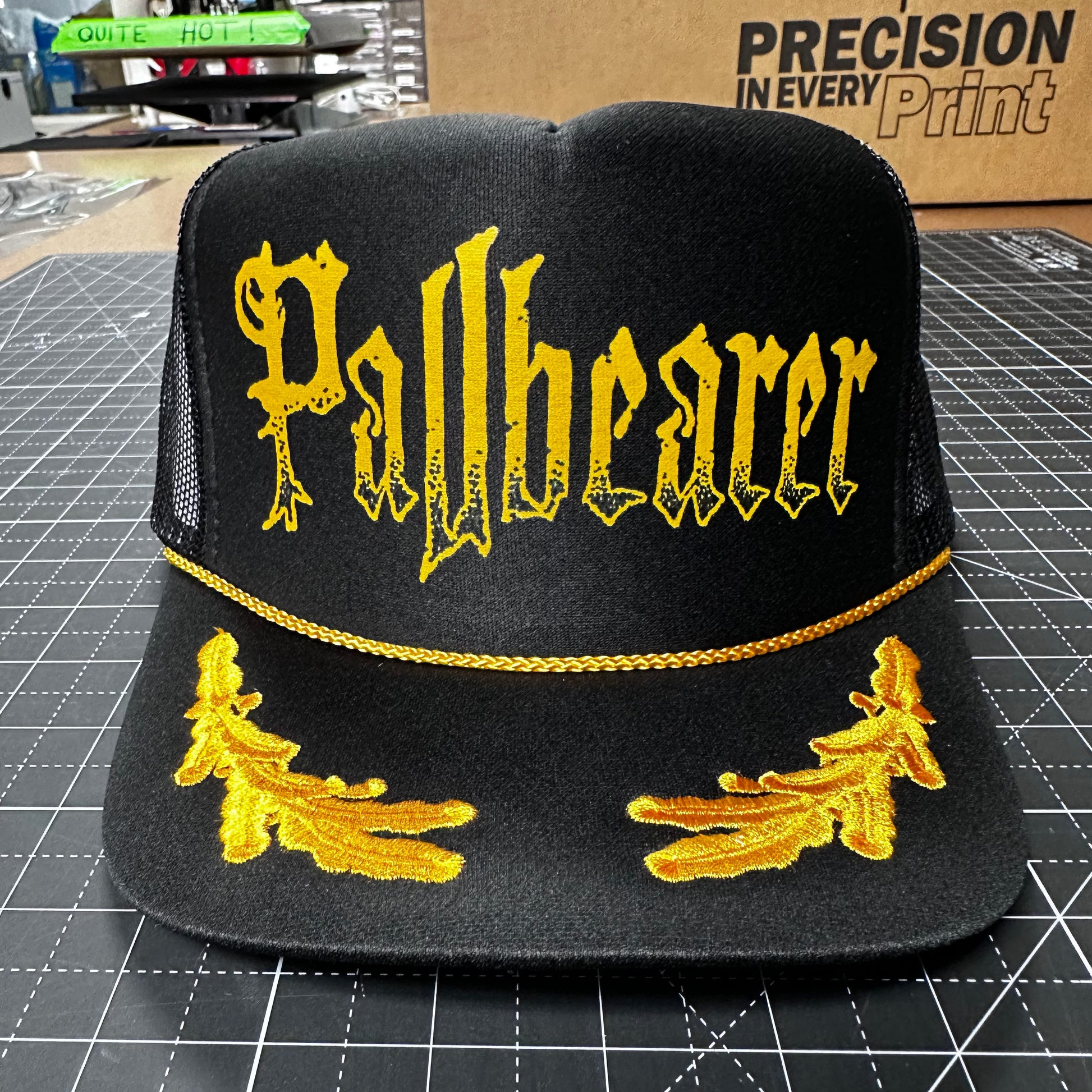 Black cap with yellow 'Pallbearer' text and designs on a grid surface.