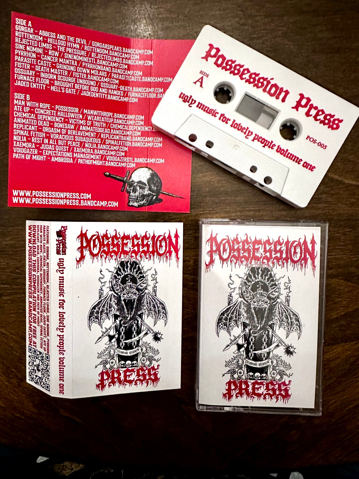 A cassette tape and its printed sleeve and insert are arranged on a dark wooden surface. The cassette is white with red text reading 'Possession Press' and 'Vol.