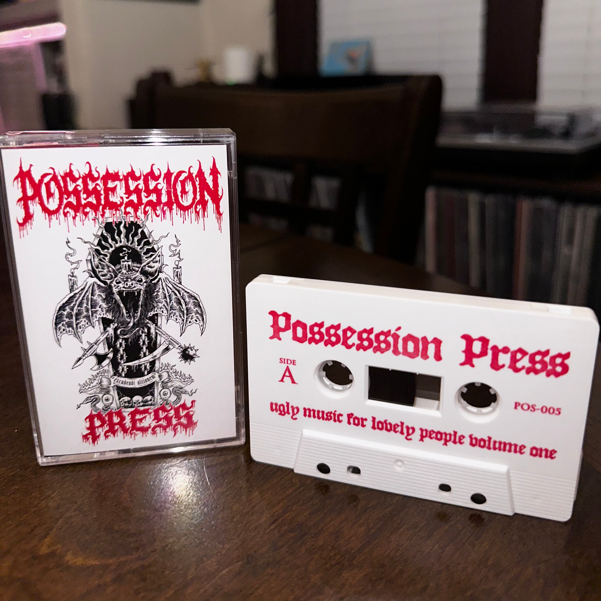 A white cassette tape and its clear jewel case cover for 'Possession Press' are displayed on a dark wooden table, with a blurred record store background.