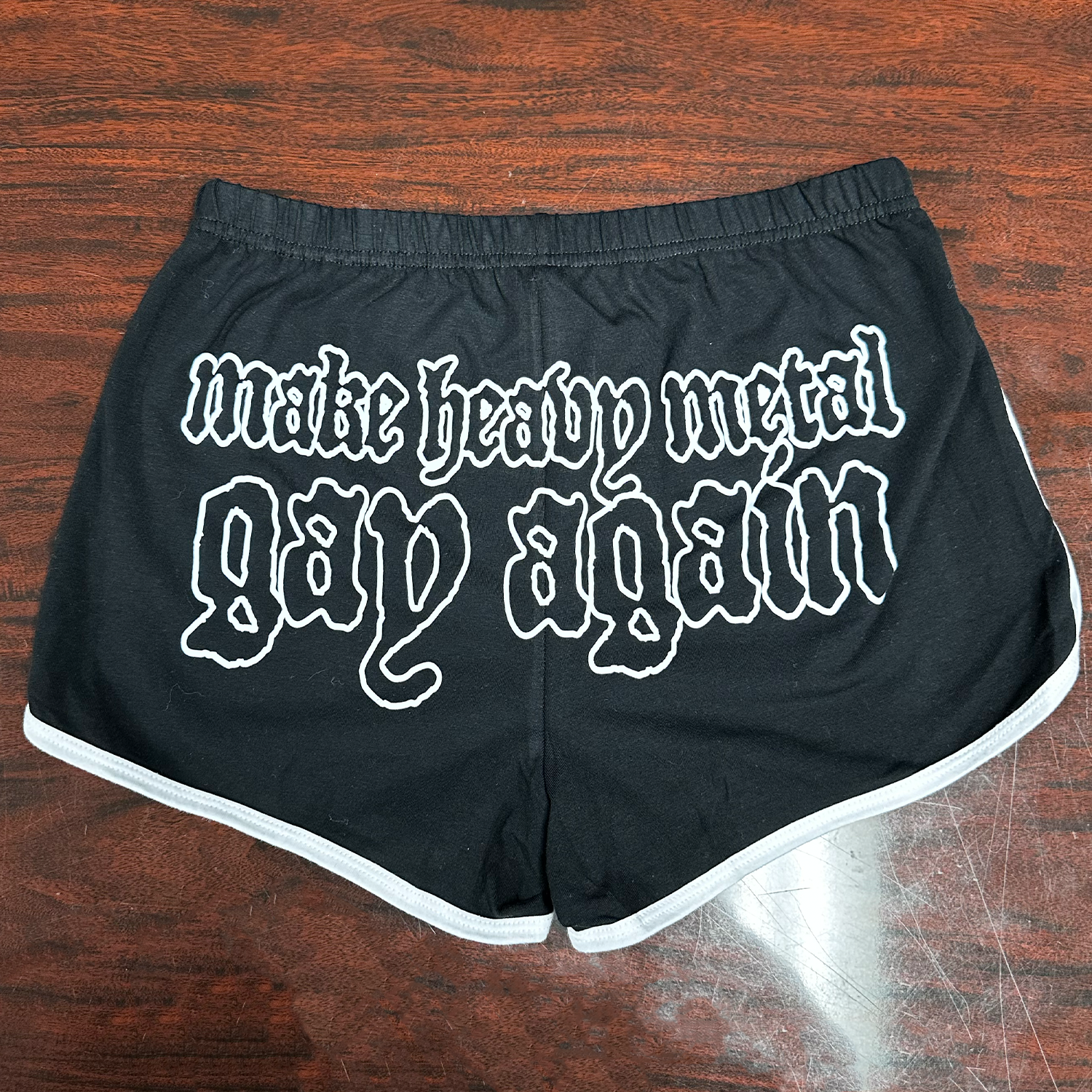 Black shorts with white "Make Heavy Metal Gay Again" text on a wooden surface