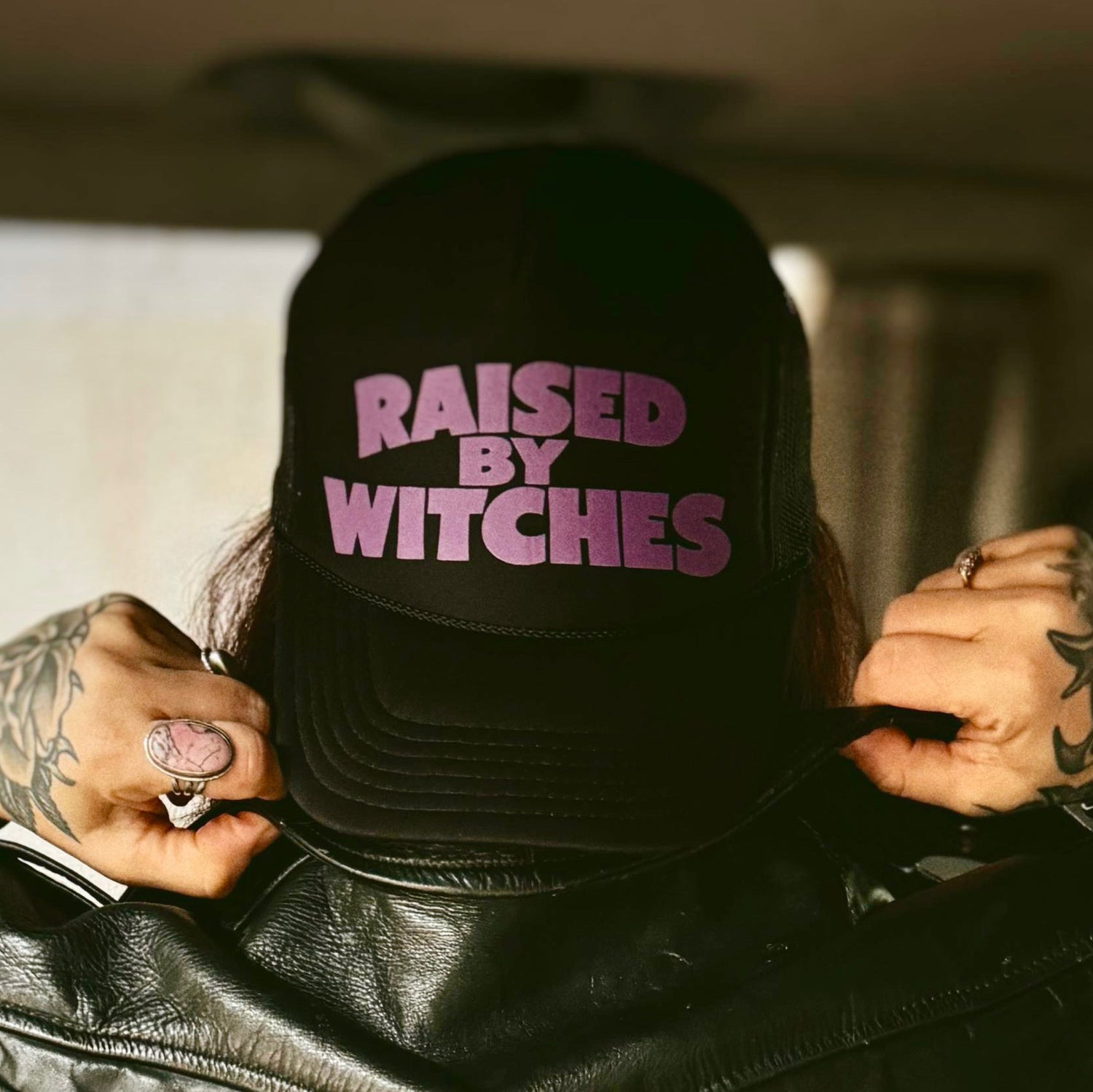 Person wearing a black cap with 'Raised by Witches' text, holding it with tattooed hands.