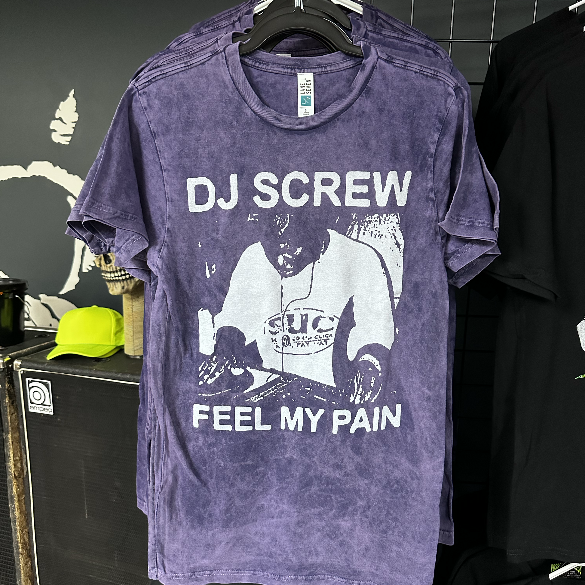 Purple t-shirt with 'DJ Screw Feel My Pain' graphic on a hanger against a dark background.